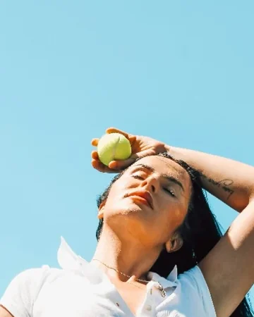 Woman in tennis whites holding a ball against a blue sky